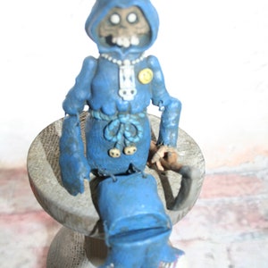 May include: A blue and gray figurine of a skeletal figure wearing a blue robe and a yellow button. The figure is sitting on a wooden base.