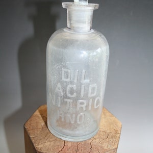 May include: A clear glass bottle with a stopper, labeled "OIL ACID NITRIC HNO3". The bottle is cylindrical with a rounded base and sits on a wooden block. The stopper is a flat disc shape.