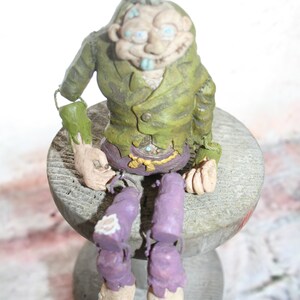 May include: A green and purple figurine of a creature with a spiky green hairdo, sitting on a wooden stool. The creature has a mischievous expression and is wearing a green jacket and purple pants.