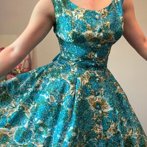 Könnte beinhalten: Ein ärmelloses Kleid mit einem lebendigen Blumenmuster in Türkis-, Petrol- und Olivgrüntönen auf weißem Grund. Das Oberteil verfügt über eine Reihe passender, stoffbezogener Knöpfe auf der Vorderseite, einen Rundhalsausschnitt und einen weiten Rock.