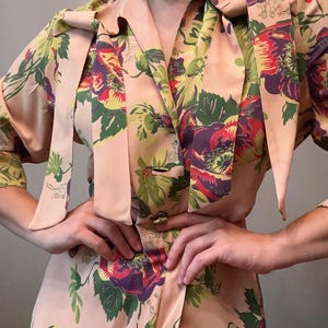 May include: A vintage dress in a soft peach colour, decorated with a vibrant floral pattern featuring red, purple, and green blooms. The dress has a collared neckline, button details, and long sleeves. The dress has a matching tie.