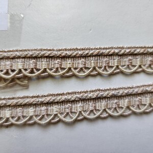 May include: Close-up of beige trim with a scalloped edge and a woven texture. The trim has a subtle shimmer and is perfect for adding a touch of elegance to any project.