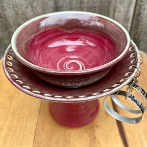 May include: A handcrafted, burgundy-colored ceramic bowl and plate set with a pedestal base. The bowl has a glossy finish with a spiral design in the center. The plate has a decorative edge. Silver hoop earrings hang from the side.