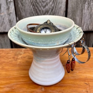 May include: A white ceramic pedestal with a light blue glaze. The pedestal has a small bowl on top and a ring for hanging earrings. A pocket watch and a pair of earrings are displayed on the pedestal.