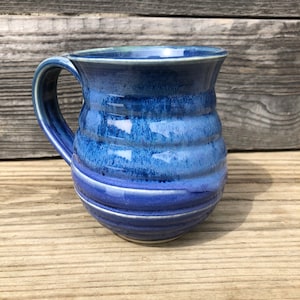 May include: A blue ceramic mug with a white rim and a textured, striped design. The mug has a rounded body and a handle.