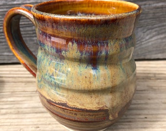 May include: A handmade ceramic mug with a brown, yellow, and blue glaze. The mug has a rounded body and a handle.