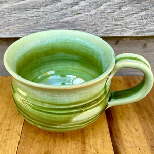 May include: A green ceramic mug with a wide, rounded bowl and a handle. The mug has a light green glaze with darker green stripes.
