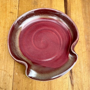 May include: A red and grey ceramic spoon rest with a wavy edge. The spoon rest has a swirl pattern.