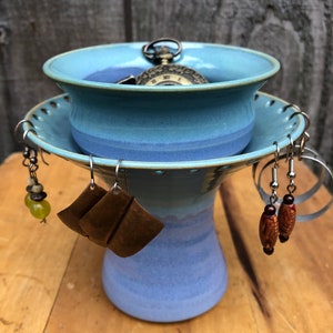 May include: A handmade ceramic jewelry holder in shades of blue. The holder has a pedestal base and a shallow bowl top. Several pairs of earrings hang from the rim, and a vintage pocket watch rests inside the bowl. The holder sits on a wooden surface.