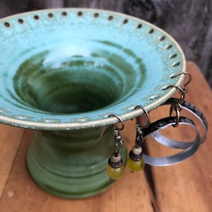 May include: A green ceramic earring holder with a wide, shallow bowl and a narrow base. The bowl has a ring of holes around the edge and two pairs of earrings are hanging from the holes. The earrings have yellow beads and silver hooks.
