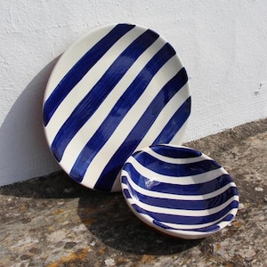 May include: Two ceramic serving pieces with a blue and white striped pattern. The larger piece is a plate, and the smaller is a bowl. The pieces are on a stone surface, with a white wall in the background.