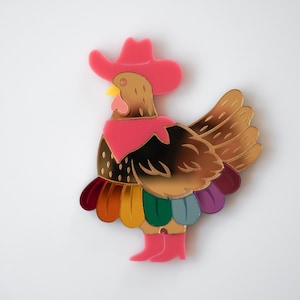 May include: A whimsical, colorful brooch in the shape of a rooster wearing a pink cowboy hat and boots. The rooster has gold and brown feathers, a yellow beak, and a pink scarf. The bottom feathers are rainbow-colored.