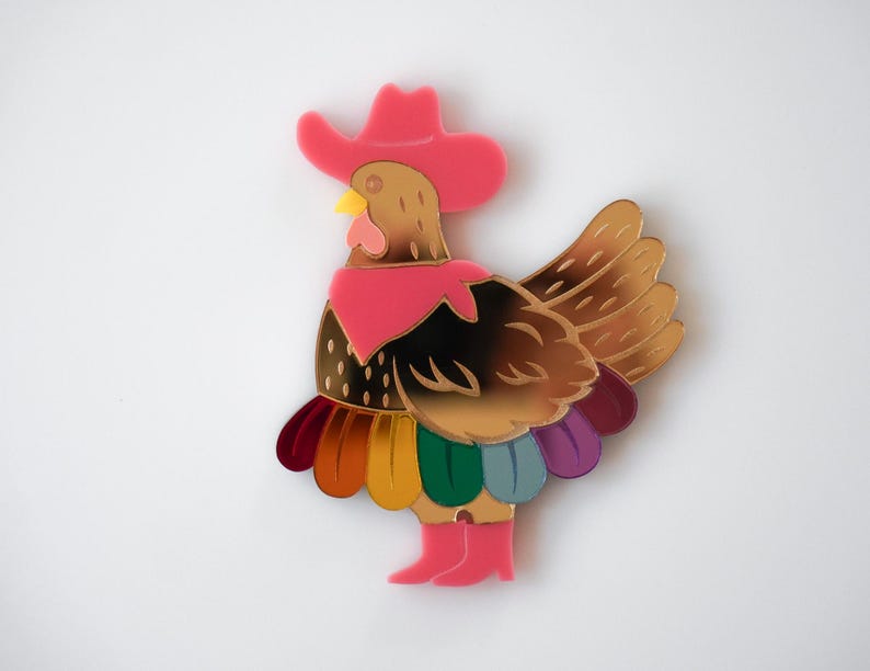 May include: A whimsical, colorful brooch in the shape of a rooster wearing a pink cowboy hat and boots. The rooster has gold and brown feathers, a yellow beak, and a pink scarf. The bottom feathers are rainbow-colored.