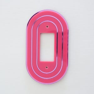 May include: A pink oval-shaped light switch cover with three concentric rings. The cover has a cut-out for a standard light switch.
