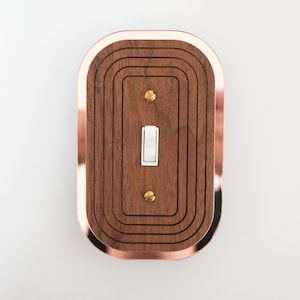 May include: A decorative light switch cover with a copper-coloured metal frame and a walnut wood centre. The wood features a series of concentric, curved grooves. The switch is white, and the cover is secured with brass screws.