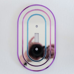 May include: A silver oval light switch cover with three concentric rings in shades of purple, pink, and blue.