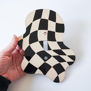 May include: A light switch cover plate with an irregular, blob-like shape featuring a black and beige checkered pattern that appears warped. The plate has a central vertical rectangular cutout for a toggle switch and two small circular screw holes.