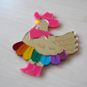May include: A whimsical, colorful acrylic rooster brooch. The rooster is gold with a pink cowboy hat, scarf, and boots. Its tail feathers are a rainbow of colors. A fun, eye-catching accessory.