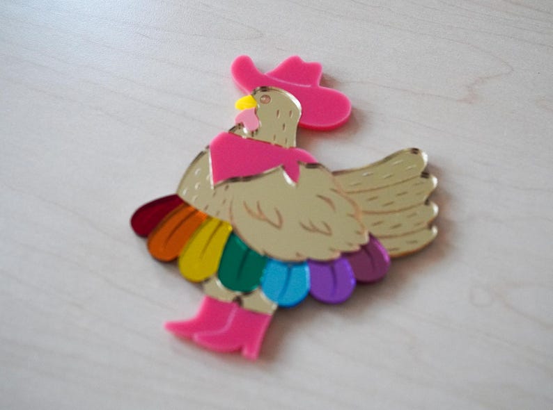 May include: A whimsical, colorful acrylic rooster brooch. The rooster is gold with a pink cowboy hat, scarf, and boots. Its tail feathers are a rainbow of colors. A fun, eye-catching accessory.