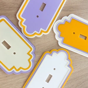 May include: Four decorative light switch covers in various colors: white, lavender, and yellow. Each cover has a scalloped edge and a rectangular opening for the switch. The covers are layered with contrasting colors.