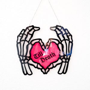 May include: A black skeleton hand-shaped wall hanging with a pink heart in the center. The heart has the words "Till Death" in black script. The wall hanging is suspended by a thin silver string.