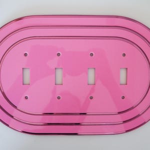 Könnte beinhalten: Eine leuchtend rosa, ovale Lichtschalterplatte mit vier rechteckigen Schalteröffnungen. Die Platte hat ein mehrschichtiges Design mit abgerundeten Kanten und kleinen Schraubenlöchern. Die glänzende Oberfläche reflektiert das Licht.