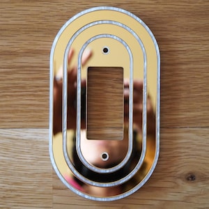May include: A gold-colored, decorative light switch cover with an oval design. The cover has three concentric oval frames, with a rectangular opening in the center for the switch. The edges are trimmed with a white, pearlescent material. Two small holes are visible.