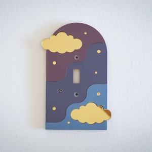 May include: A purple and blue light switch cover with a gold cloud design. The cover has a rounded top and a cutout for a light switch.