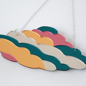 May include: A colorful cloud-shaped wall hanging. The layered design features shades of gold, teal, pink, and cream. A silver chain is attached for hanging. The decorative piece is a modern art object.