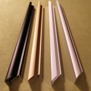 May include: Four picture frame moulding samples in black, beige, white, and off-white. The samples are angled and have a channel for holding a picture. The mouldings are arranged on a green surface.