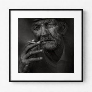 Op de afbeelding: Zwart-wit ingelijste print van een oudere persoon met een snor en een hoed, die een brandende sigaret vasthoudt. Het portret is een close-up, gericht op het gezicht en de handen van de persoon. De afbeelding is in grijstinten, met een focus op de details van de gelaatstrekken.