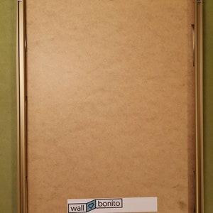 May include: A gold-coloured picture frame with a brown backing. The frame has a hanging hook on the top and a sticker on the bottom that says "wall &hearts; bonito".