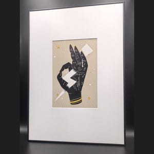 Può includere: Una stampa incorniciata con l'illustrazione di una mano nera su sfondo beige. La mano presenta simboli e linee astrologiche bianche e tiene in mano una forma di fulmine bianco. Piccole stelle dorate circondano la mano, il tutto racchiuso in un passe-partout bianco e una sottile cornice nera.