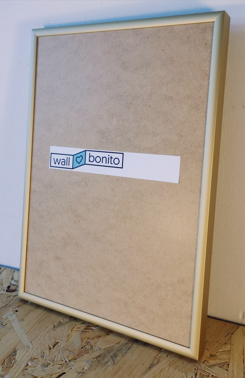 May include: A gold-colored picture frame with a light brown backing. The frame has a sticker on the front that says "wall &hearts; bonito".