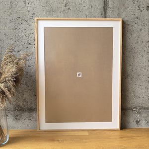 Può includere: Una cornice rettangolare in legno con un passe-partout bianco e un retro marrone. La cornice è vuota, con un piccolo adesivo quadrato al centro. La cornice è esposta su una superficie di legno contro un muro di cemento grigio.
