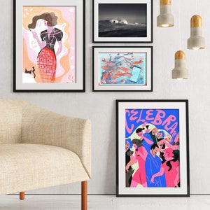 May include: Four framed illustrations on a white wall. The top left illustration is a pink and red illustration of a woman in a black dress. The top right illustration is a black and white photograph of a wave crashing in the ocean. The bottom left illustration is a colorful illustration of fish swimming in a pond. The bottom right illustration is a colorful illustration of people dancing with the text "CELEBRATE" in blue.