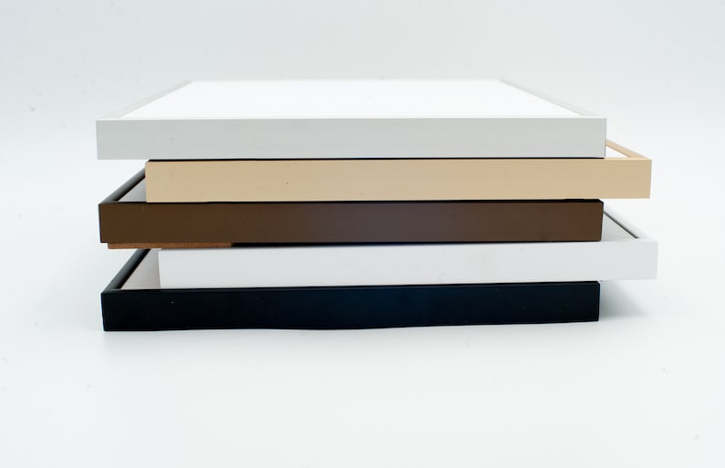 May include: A stack of five square picture frames in various colors. The frames are in white, light gray, beige, brown, and black. The frames are stacked on top of each other, with the white frame on top.