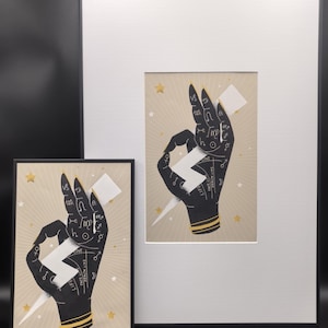 May include: Two framed art prints featuring a black hand with gold accents, holding a white lightning bolt. The hand is adorned with astrological symbols and lines. The background is beige with gold stars. One print is smaller, in a black frame.