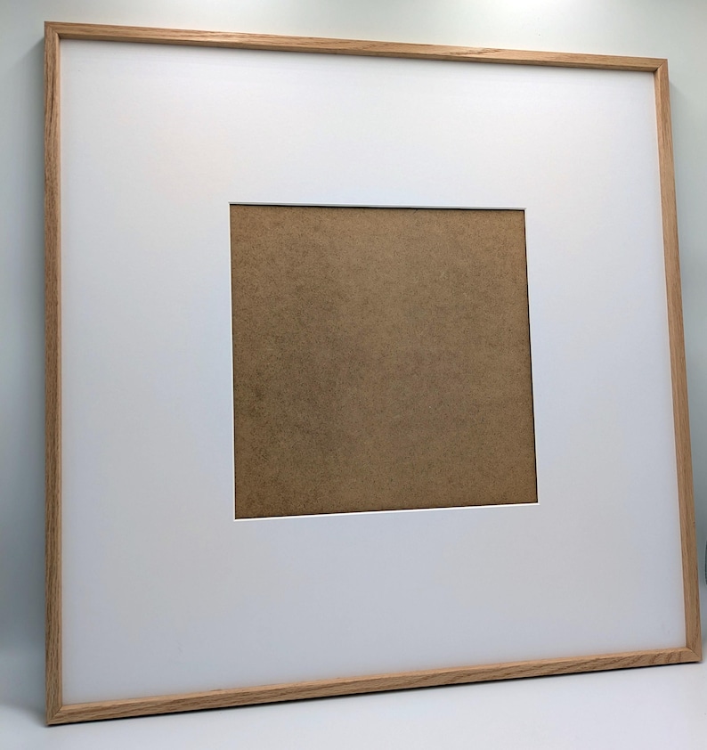 May include: Square picture frame with a light wood grain finish. The frame has a white mat with a square opening, revealing a brown backing. The frame is designed for displaying artwork or photographs.