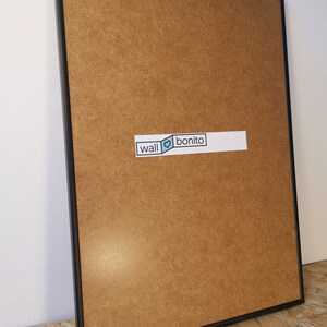 May include: A black framed picture with a brown wood backing. The frame is on a wooden surface. A sticker on the backing says "wall &hearts; bonito".