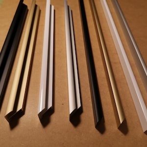 May include: Eight different picture frame molding samples in various colors and finishes. The samples are arranged in a row from left to right, with the black molding on the left and the white molding on the right. The molding is made of metal and has a smooth, polished finish.
