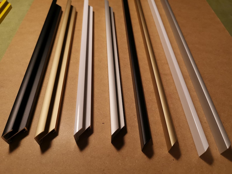 May include: Eight different picture frame molding samples in various colors and finishes. The samples are arranged in a row from left to right, with the black molding on the left and the white molding on the right. The molding is made of metal and has a smooth, polished finish.