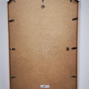 Op de afbeelding: De achterkant van een lichtbruin houten fotolijst met een wit label waarop "wall bonito" staat. De lijst heeft metalen ophangbeugels aan de achterkant.