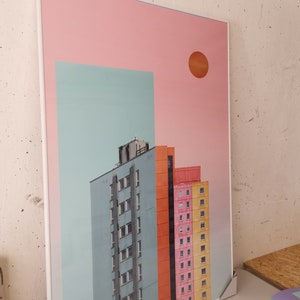 K&ouml;nnte beinhalten: Ein gerahmter Druck eines Stadtbildes mit drei Geb&auml;uden in verschiedenen Farben: blau, orange und rosa. Die Geb&auml;ude stehen vor einem hellblauen Himmel mit einer gro&szlig;en orangefarbenen Sonne oben rechts. Der Hintergrund ist hellrosa.