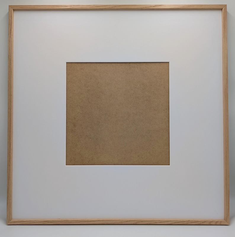 May include: Square wooden picture frame with a white mat and a brown backing. The frame is made of light-colored wood and has a simple, modern design. The mat opening is square, and the backing is a solid brown color.