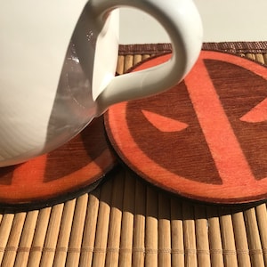 May include: Two round wooden coasters with a red and brown Deadpool logo. The coasters are on a bamboo mat.