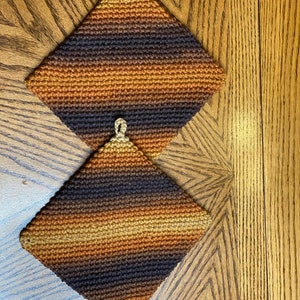 May include: Two crocheted pot holders in a brown, tan, and black striped pattern. The pot holders have a loop at the top for hanging.