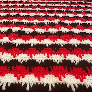 May include: A crocheted blanket with alternating stripes of brown, white, and red yarn. The blanket has a textured, fuzzy look.