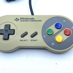 May include: A vintage Nintendo Super Famicom controller. The controller is a light gray color with a gray circular button area. It has a directional pad, select and start buttons, and colored action buttons: blue, green, red, and yellow.