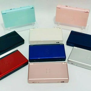 May include: A collection of Nintendo DS handheld gaming consoles in various colors, including mint green, pink, black, white, blue, silver, red, and rose gold. The consoles are displayed on clear stands, showcasing their design.